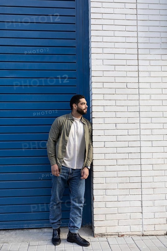A man in casual attire leans against a white brick wall of the city, with a blue door beside him. He is wearing jeans, a white T shirt, and a green jacket, exuding a relaxed and stylish vibe