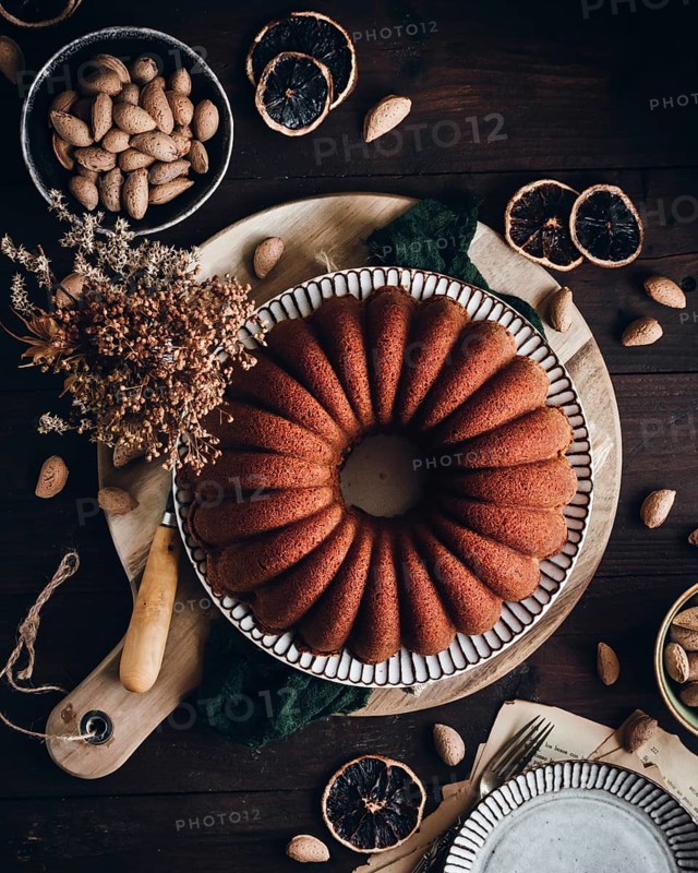 A beautifully baked bundt cake infused with AOVE, almond, and orange sits on a rustic wooden table, surrounded by whole almonds, dried orange slices, and decorative elements