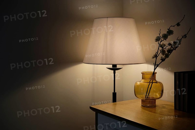 Cozy home interior detail. Warm light from a floor lamp illuminates a corner with an amber glass vase, flowers, and books on a wooden surface