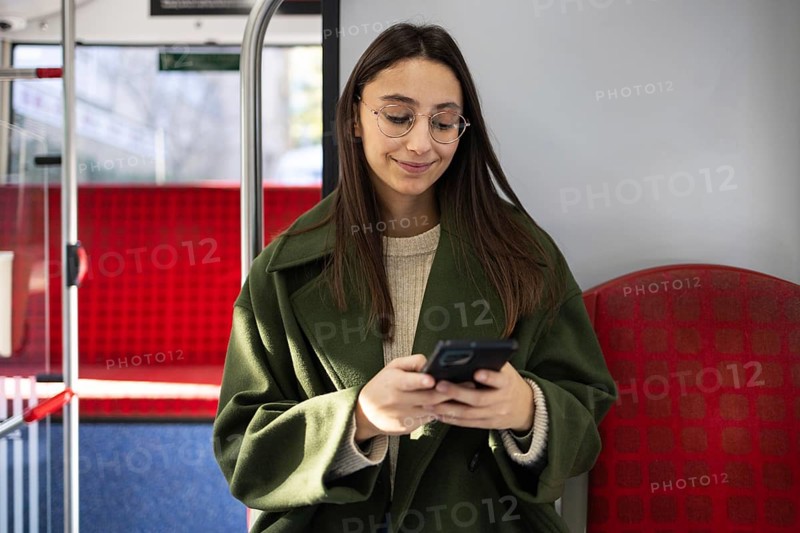A woman in a green coat, with glasses, is using her smartphone while seated on public transport Natural light highlights her relaxed expression in this urban setting