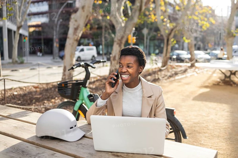 A black woman engages in a lively phone call while sitting at a wooden table with her laptop in a vibrant city park, exuding confidence and joy in an urban setting