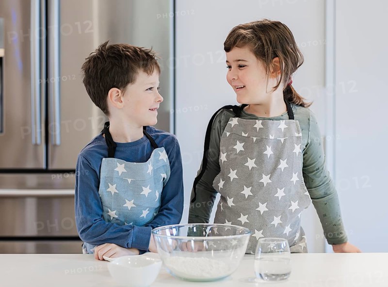Two children wearing matching star patterned aprons stand at a kitchen counter, smiling at each other. A bowl with ingredients and a glass of water are nearby
