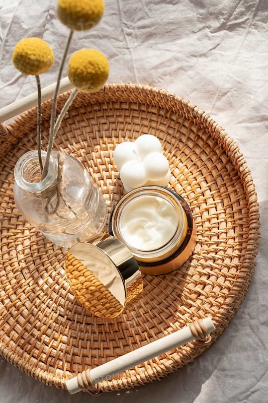 A cosmetic of skincare cream, a decorative candle, and a vase with yellow flowers rest on a woven tray, capturing a serene and natural beauty ambiance