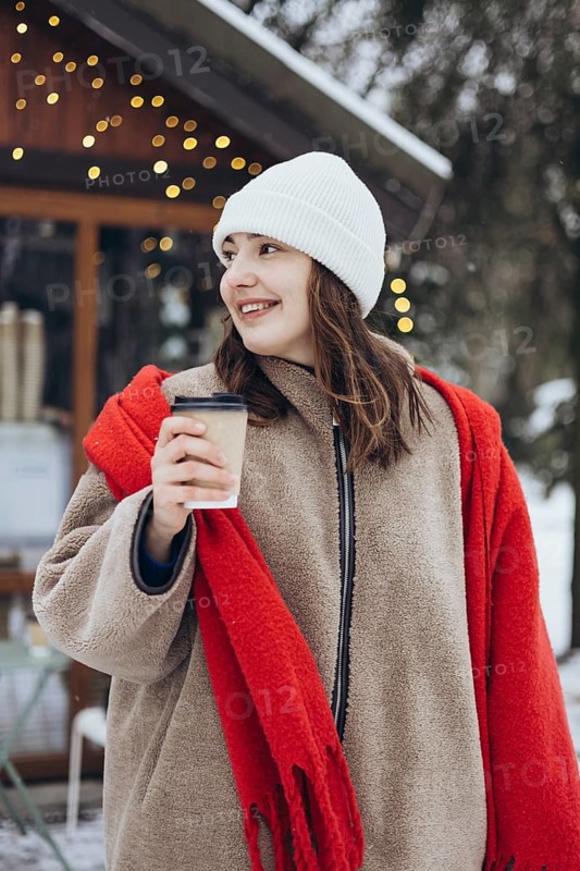 A young woman smiles gently as she holds a cup of coffee outdoors during a snowy winter day. She is wearing a white beanie, red scarf, and a cozy beige coat with festive lights in the background