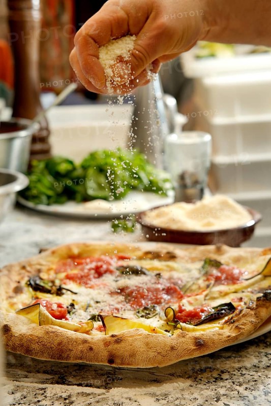 Cropped unrecognizable person sprinkling grated cheese on a freshly baked pizza topped with vegetables and sauce in a stylish kitchen setting