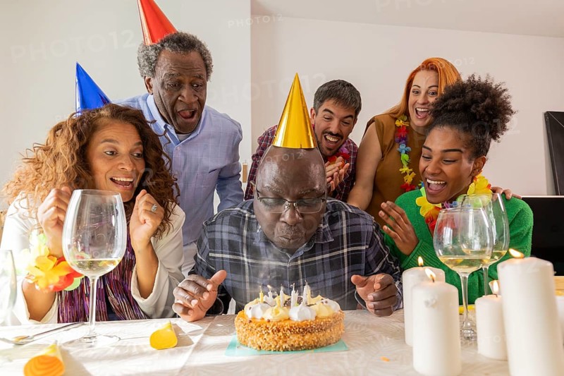 A joyful group at a festive birthday celebration. The guest of honor wears a party hat, ready to blow out candles on a cake, surrounded by friends sharing in the joy