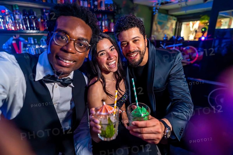 Cheerful multi ethnic friends taking selfie and showing cocktails while enjoying nightlife at nightclub bar