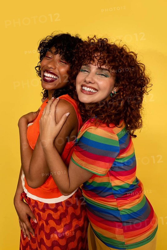 Two joyful women embrace warmly, expressing happiness and friendship. They wear bright, colorful clothing against a vibrant yellow background, highlighting a festive atmosphere