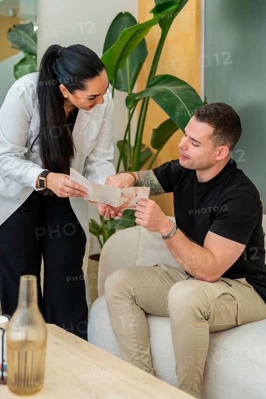 Doctor in white lab coat explaining aesthetic treatment options to a male patient seated on a sofa in a modern clinic, discussing skincare, procedures and personalized care