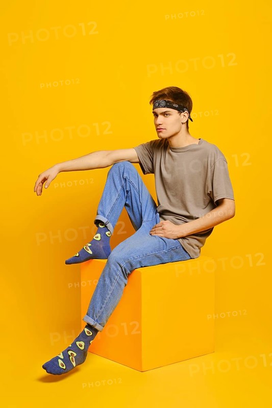 Young man sitting on cube over yellow studio background
