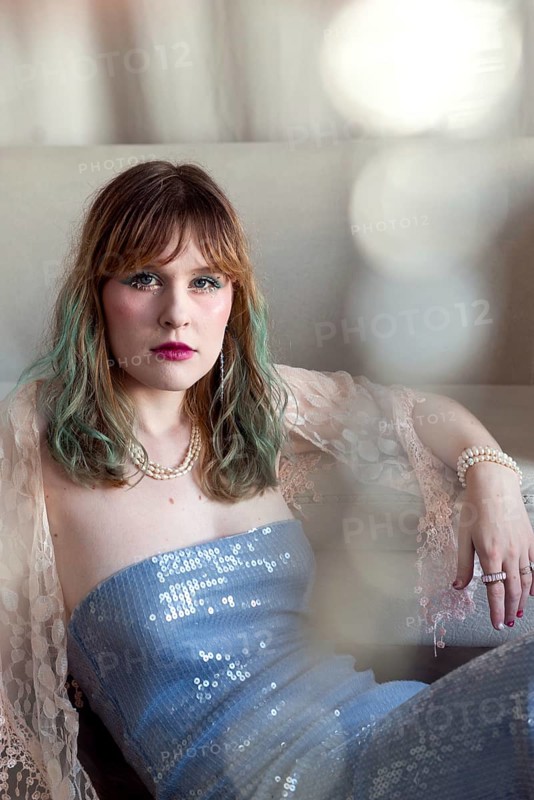 A woman in a blue sequin dress, highlighted by pearls and lace, reclines elegantly on a sofa Her gaze is confident, reflecting glam and sophistication