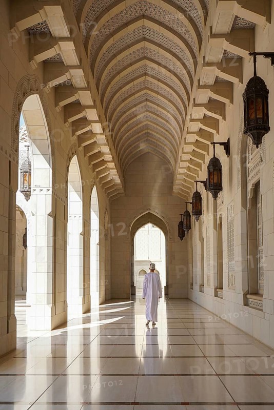 Back view of unrecognizable man in traditional dishdasha Omani attire walkoing along a corridor inside the Sultan Qaboos Mosque in Muscat, Oman, showcasing the intricate Islamic architectural and design details and hanging lanterns