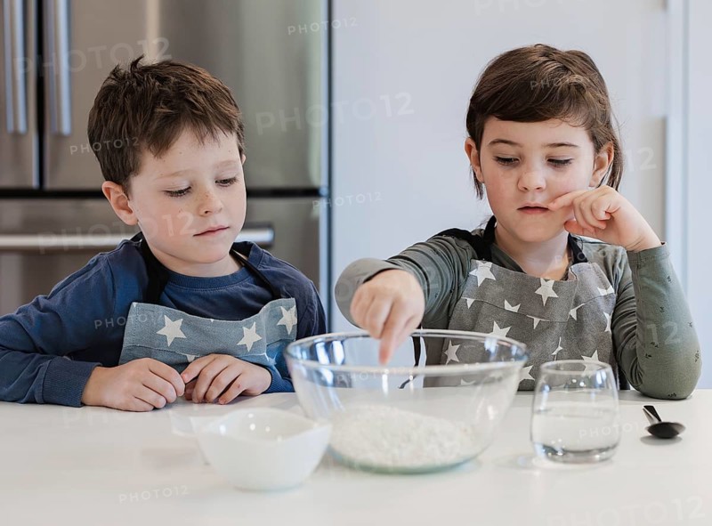 Two siblings wearing star patterned aprons are engaged in baking activities in a modern kitchen. One child is reaching into a mixing bowl with flour while the other observes attentively