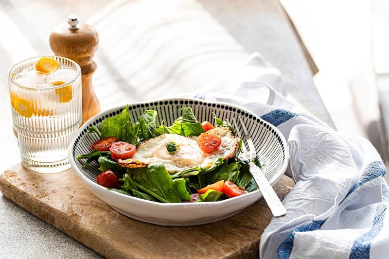 Top view of a healthy lunch featuring fresh spinach leaves, cherry tomatoes, and a fried egg in a stylish bowl, accompanied by a glass of mineral water with ice and kumquat slices