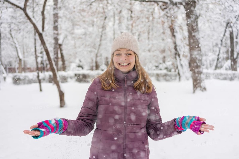 A woman in a purple jacket and knit hat smiles joyfully while standing in a snowy park. The winter scene is filled with snow-covered trees and a serene atmosphere