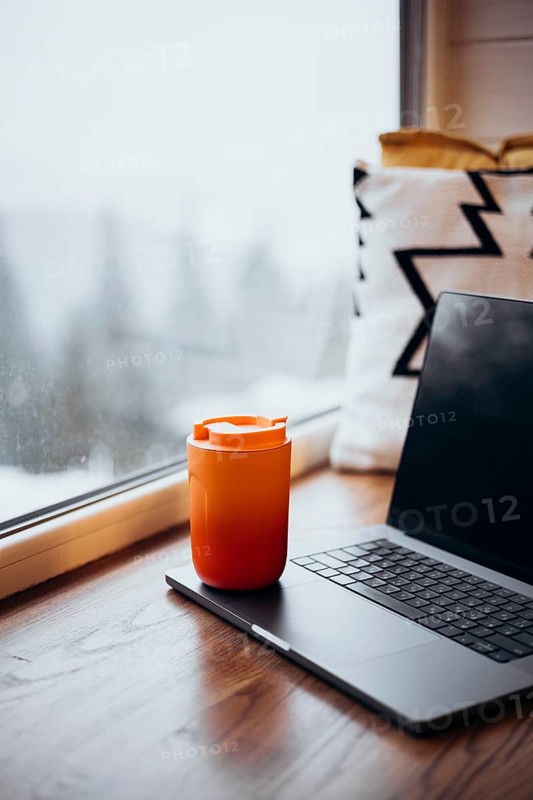 A laptop beside a vibrant orange thermos cup set on a wooden table, illustrating a comfortable remote work environment. The setup overlooks a frosty window, adding a wintry atmosphere to a modern work-from-home scene