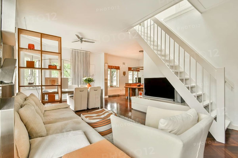Spacious open concept living room featuring cozy cream sofas, a sleek staircase, and large windows. The elegant decor includes a dining area, modern shelving, and a stylish ceiling fan
