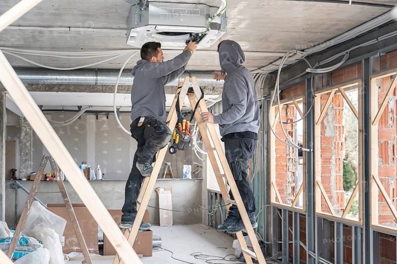 Air conditioning technicians are installing an hvac unit on the ceiling of a new building, standing on ladders and connecting wires in a renovation and construction environment