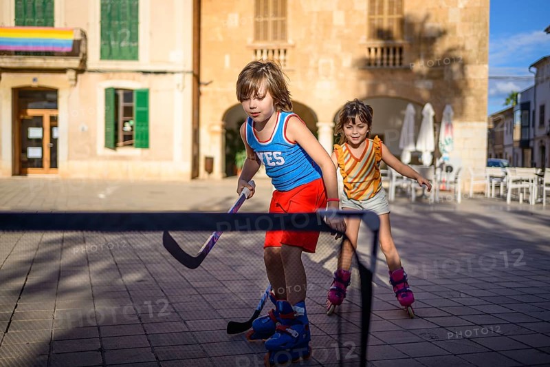 Two children play street hockey on roller blades in a sunlit square. They are focused and energetic, surrounded by charming architecture and warm afternoon light