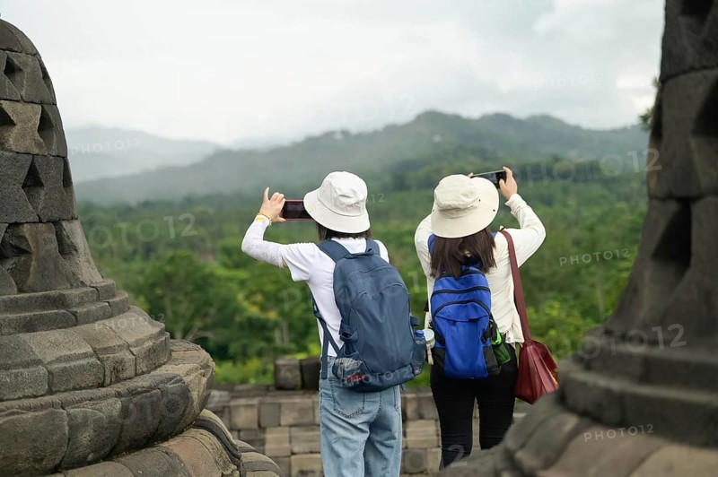 Back view of unrecognizable tourists wearing hats and backpacks, standing at the historic Borobudur Temple. They are photographed capturing the lush green landscape and distant hills using a smartphone and a camera