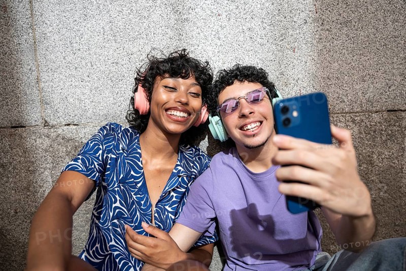 A joyful moment between a multiethnic LGBTQ+ couple as they take a selfie outdoors, wearing colorful headphones and casual outfits, capturing their happiness and love