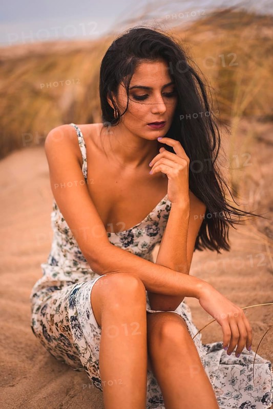Portrait of a brunette Caucasian caressing her hair in a white floral dress, sitting on the sand on the beach of Cabo de Gata, Nijar. Andalucia, Spain