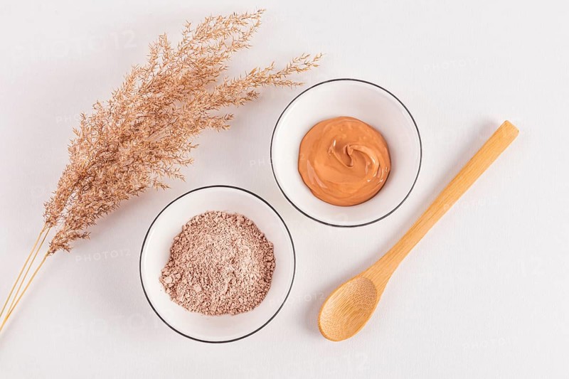 Top view of pink cosmetic clay powder and prepared facial mask in bowls with a decorative grass twig on a white surface, depicted in a minimalistic style