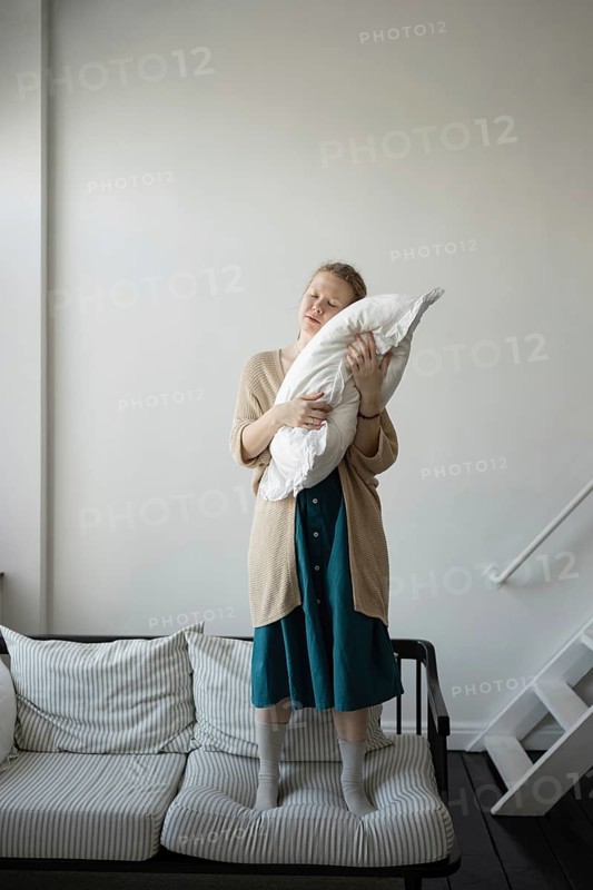 A woman stands on a couch in a cozy living room, embracing a pillow with her eyes closed, conveying a sense of relaxation and comfort in a peaceful home environment