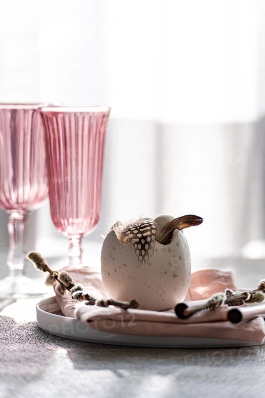 A stylish Easter dining setup with a focal decorative egg adorned with feathers, nestled on a pastel tablecloth, complemented by elegant pink-tinted glasses