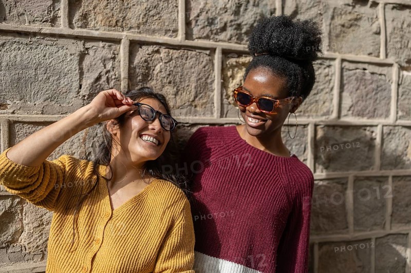 Two friends are laughing and enjoying a sunny day while wearing trendy sunglasses, standing against a textured stone wall. Their casual attire and joyful expressions create a vibrant mood