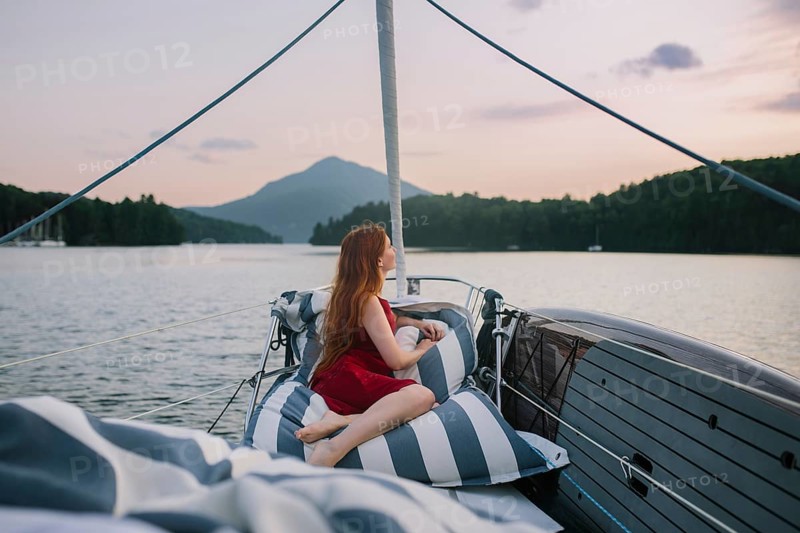 A person in a red dress relaxes on cushions aboard a sailboat, enjoying a peaceful, scenic afternoon on the water with a distant mountain view under a soft pink sky
