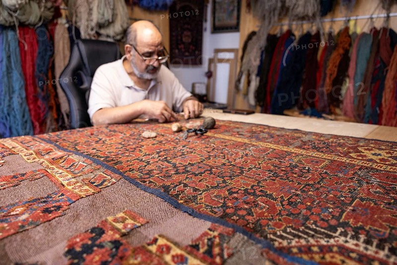 A senior senior male Iranian carpet restorer carefully repairs a traditional rug in his Salzburg workshop, surrounded by textile materials