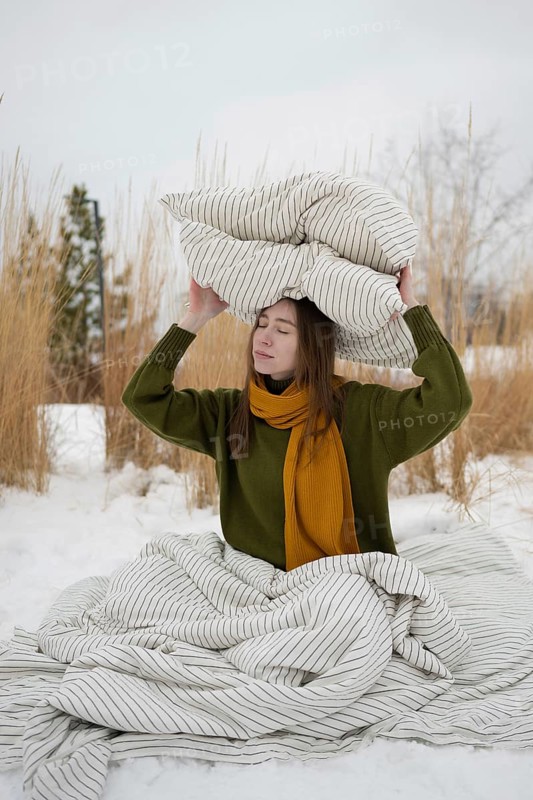 A woman sits on snow, wrapped in a striped blanket, holding a pillow on their head. Wearing a green sweater and yellow scarf, they exude warmth in a snowy park