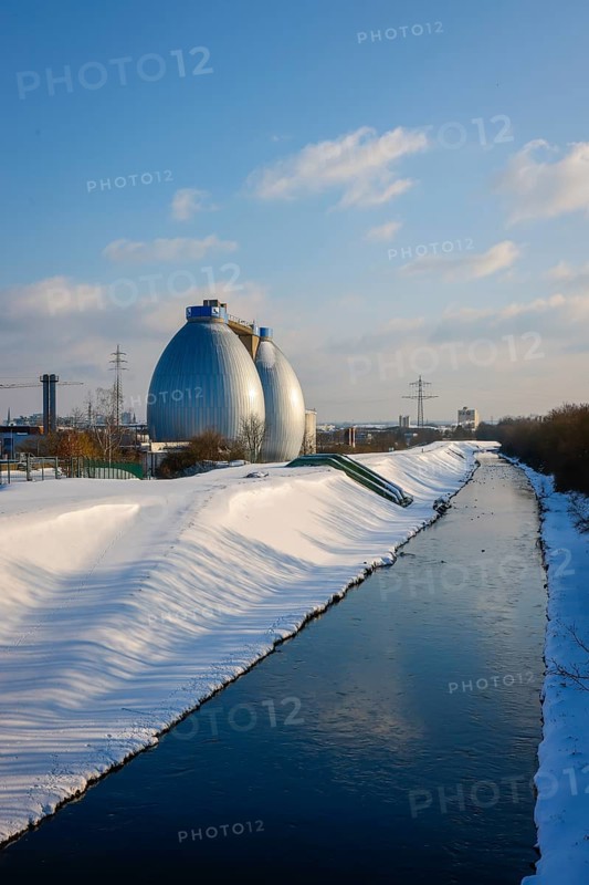 Dortmund, North Rhine-Westphalia, Germany - Sunny winter landscape in the Ruhr area, ice and snow on the renaturalised Emscher, digestion towers at the Dortmund Deusen sewage treatment plant, the treatment plant is now being equipped with a 4th purification stage, which also removes drug residues from the treated wastewater, the ecological conversion of the Emscher system consists of the construction of a central wastewater treatment system in the Ruhr area, the construction of underground sewers and the renaturation of the Emscher and its tributaries