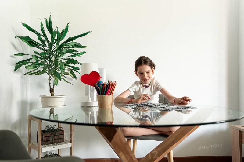 A disabled girl with a tracheostomy sits at a table, focusing on a puzzle. Surrounded by art supplies and a lamp with a heart, she appears happy and engaged in her activity