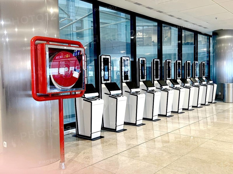 A row of self-service kiosks in a modern airport setting, alongside a fire safety hose reel. The scene reflects efficiency and safety in air travel environments