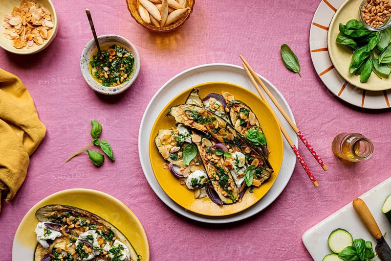 Top view of a gourmet dish featuring roasted courgettes topped with mozzarella, basil, and toasted almonds beautifully arranged in a yellow plate, with dressing and pine nuts on the side, laid on a textured pink tabletop