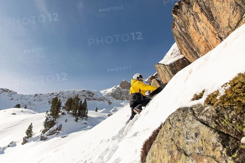 A snowboarder in a vibrant yellow jacket climbs a snow-covered slope in the backcountry. The scene captures the thrill of freeride snowboarding amidst majestic winter mountains