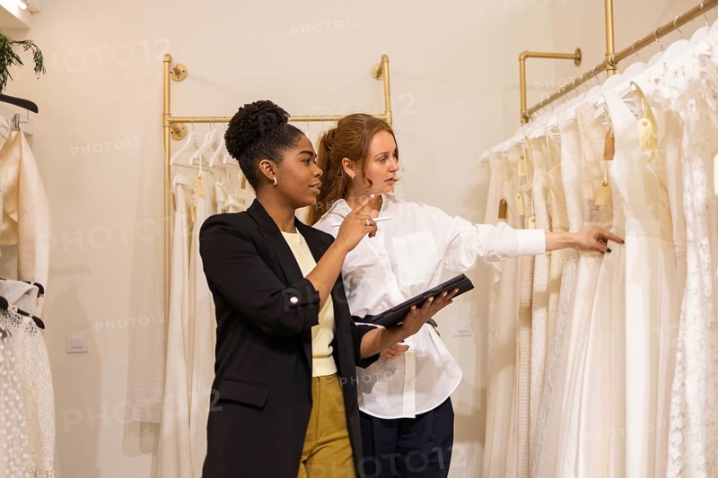 Two women explore a bridal boutique, deciding on wedding dresses. One holds a tablet for details, while the other points at gowns, highlighting a moment of fashion decision making