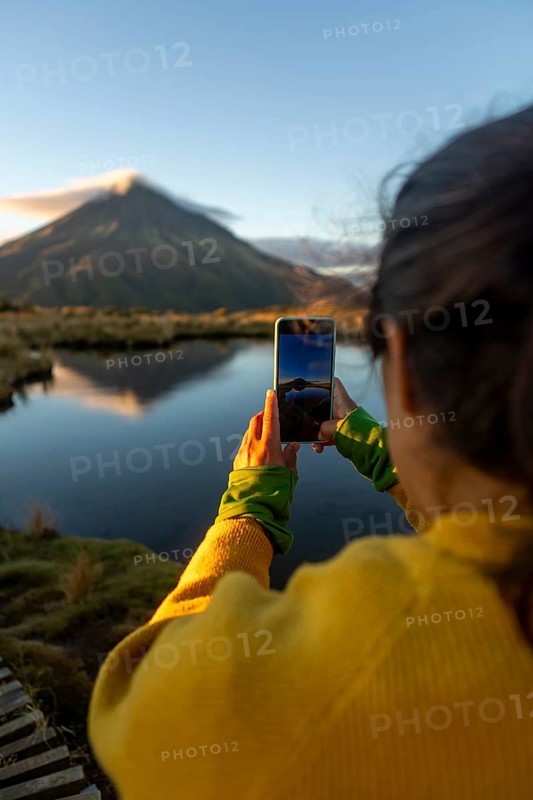 Back view of cropped unrecognizable female traveler captures the stunning sunrise view of Mount Taranaki in New Zealand, reflecting on a calm lake, using her smartphone during an outdoor adventure