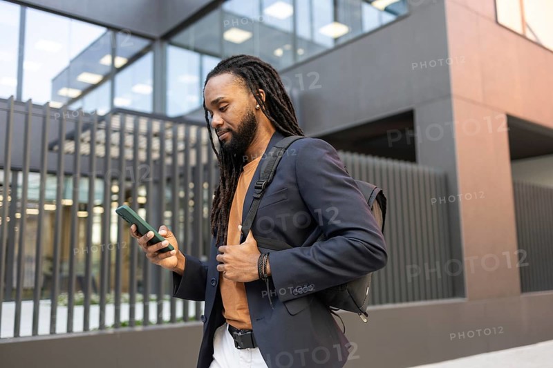 Business man wearing a blazer and backpack, walking outside a modern office building while checking his smartphone. Perfect for themes of business, mobility, or technology