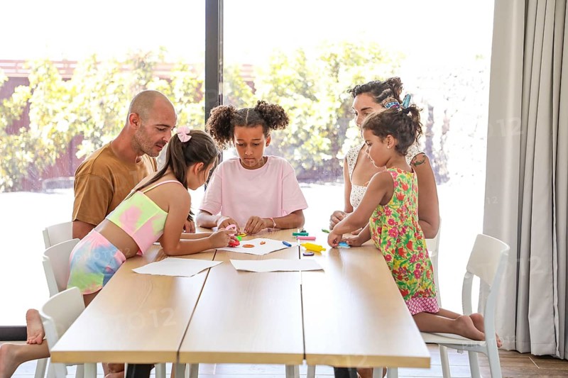 A family gathers around a wooden table, engaging in creative activities together. The bright room is filled with natural light, highlighting the joyful interaction and shared moments
