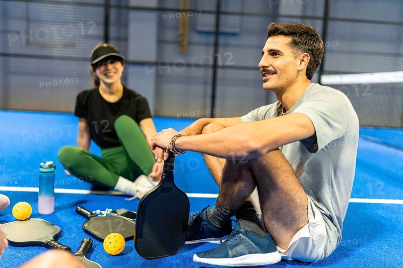 Two friends resting on a blue pickleball court, holding paddles and listening, enjoying a moment of camaraderie and strategy during their athletic activity