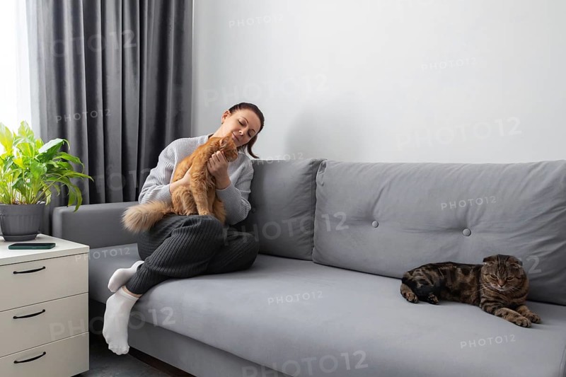 A relaxed woman cuddles her ginger cat on a gray couch, while another cat lounges beside her in a cozy home setting with neutral decor and a touch of greenery