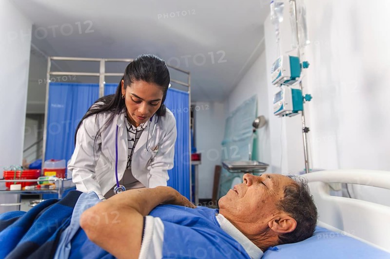 A female doctor attentively examines an elderly male patient in a hospital setting. The room is equipped with medical devices, emphasizing a caring healthcare environment