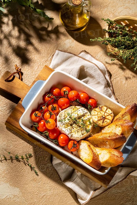 Savor the sight of baked Camembert with cherry tomatoes, garlic, and fresh herbs. The dish is completed with toasted baguette slices, offering a delightful culinary experience