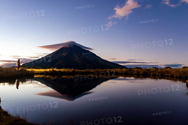 Side view of unrecognizable person admiring the tranquil reflection of Mount Taranaki in a serene lake during sunrise in Nueva Zelanda, capturing a majestic landscape