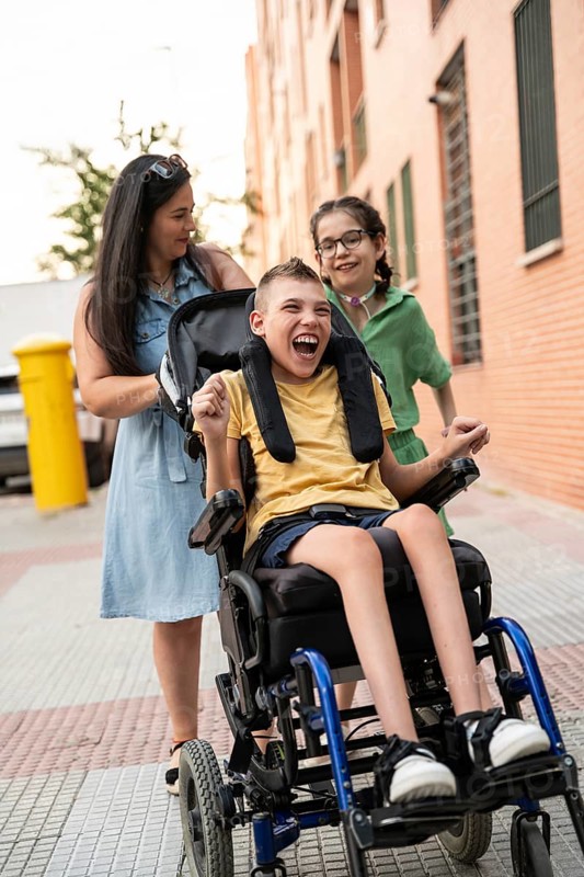 A family enjoys a day out. The mother smiles, pushing her son, who has cerebral palsy, in a wheelchair. Beside them is the daughter, a girl with a tracheostomy, who is smiling too