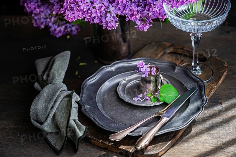 Elegant table setting with vintage plates, delicate lilac flowers in a tin cup, and a graceful glass perched on a wooden board, exuding rustic charm and warmth