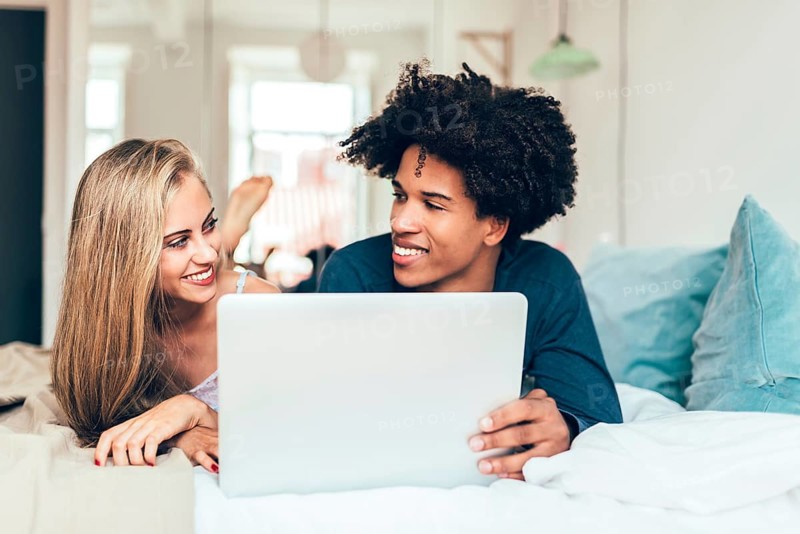 A cheerful couple lies on their bed, engaged with a laptop. The cozy bedroom setting and their shared laughter create a warm, affectionate atmosphere perfect for bonding